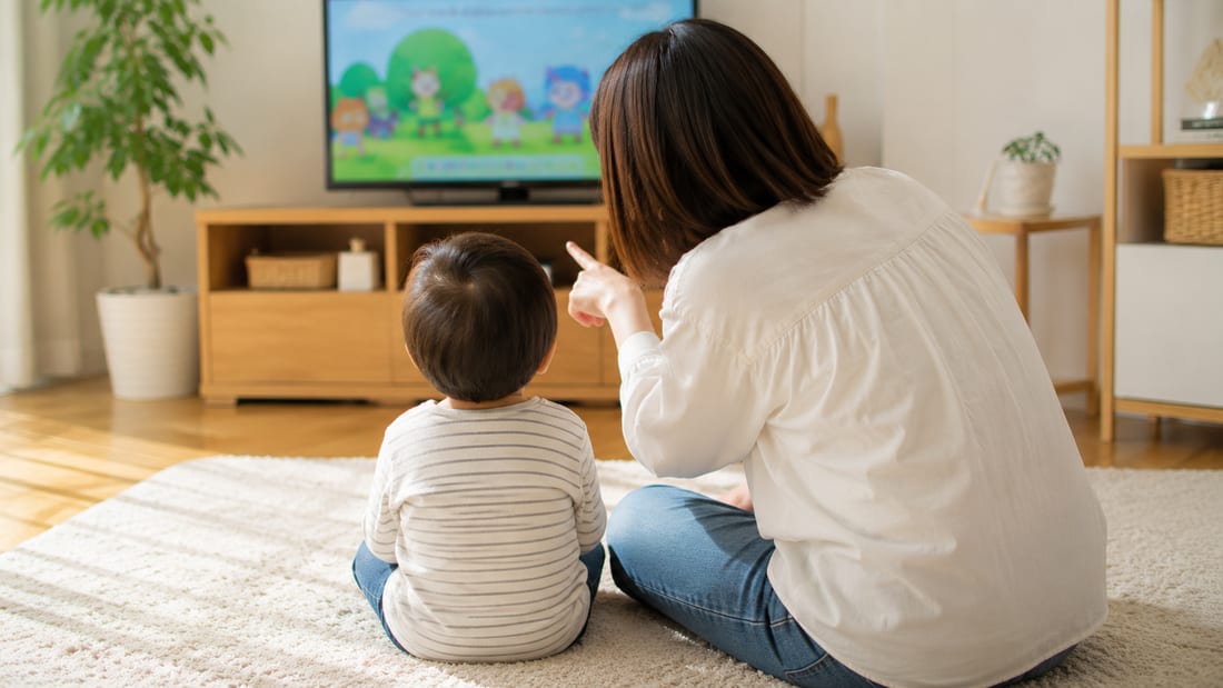 リビングでテレビを見る子どもの後ろ姿と、横から声をかける親の手元
