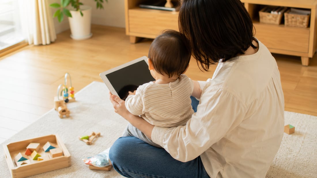親のひざの上に座ってタブレットを見る小さな子どもの後ろ姿